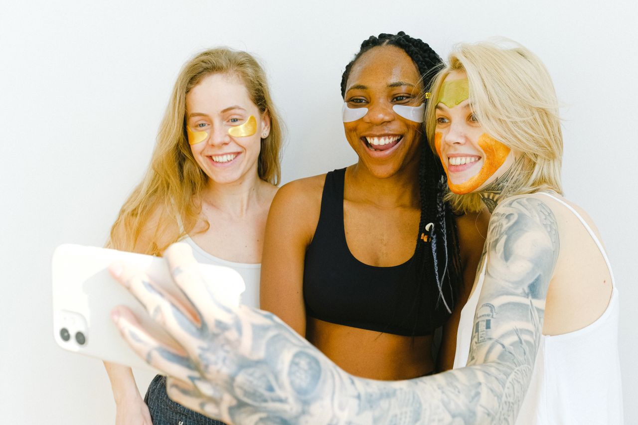 töitämme Group of happy women laughing while having fun with cosmetic eyes patches and mask for face while standing together and taking selfie on smartphone