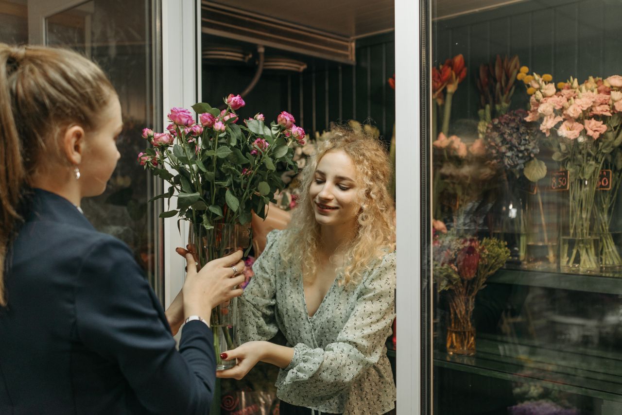 töitämme Two women exchanging a bouquet in a charming flower shop setting.